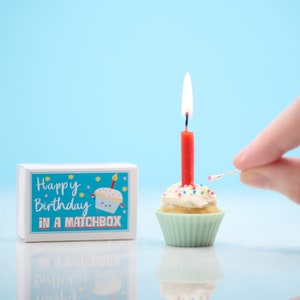 May include: A small white cupcake with colourful sprinkles and a red candle is lit with a match. The cupcake is sitting on a blue background. A white matchbox with the text "Happy Birthday in a Matchbox" is in the background.