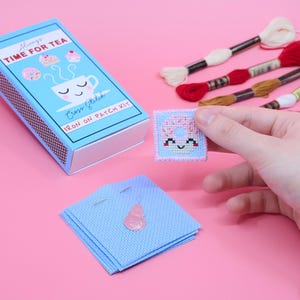 May include: A blue matchbox-style box with the text "Always Time for Tea" and "Cross Stitch Iron On Patch Kit". The box contains a small cross-stitch patch of a pink donut with a smiling face.