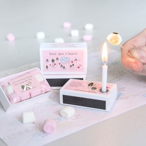 May include: A s'mores gift set with a matchbox candle holder, marshmallows, and matchboxes with text. The marshmallows are pink and white. A marshmallow is being toasted over a lit candle. The text reads "love you s'more".