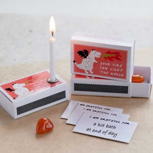 May include: A set of three matchbox-style boxes with a dragon illustration and the text "Your fire can light the world". Each box contains a small candle, a crystal, and a card with the text "I am grateful for..." followed by a different phrase on each card.