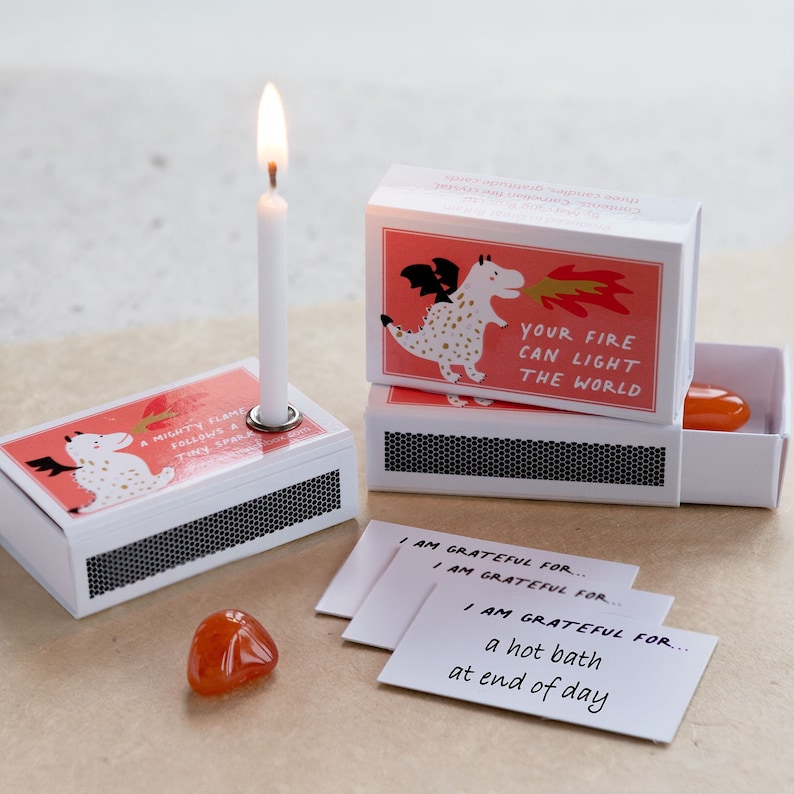 May include: A set of three matchbox-style boxes with a dragon illustration and the text "Your fire can light the world". Each box contains a small candle, a crystal, and a card with the text "I am grateful for..." followed by a different phrase on each card.