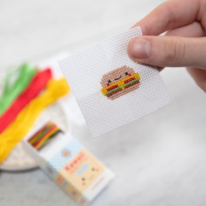May include: A white cross-stitch embroidery of a cartoon burger with a happy face. The burger is made with yellow cheese, green lettuce, and red tomato.