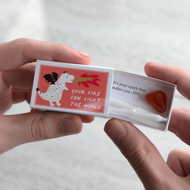 May include: A white matchbox with a red and white illustration of a dragon blowing fire. The text on the box reads "Your fire can light the world." The box is open and contains a small orange crystal and a white candle.