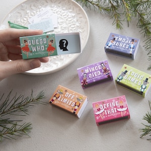 May include: A collection of matchbox games, including "Guess Who," "Emoji Tunes," and "Logic Games." Each box features colorful illustrations and text, with a festive theme. The boxes are arranged on a white plate with sprigs of greenery.