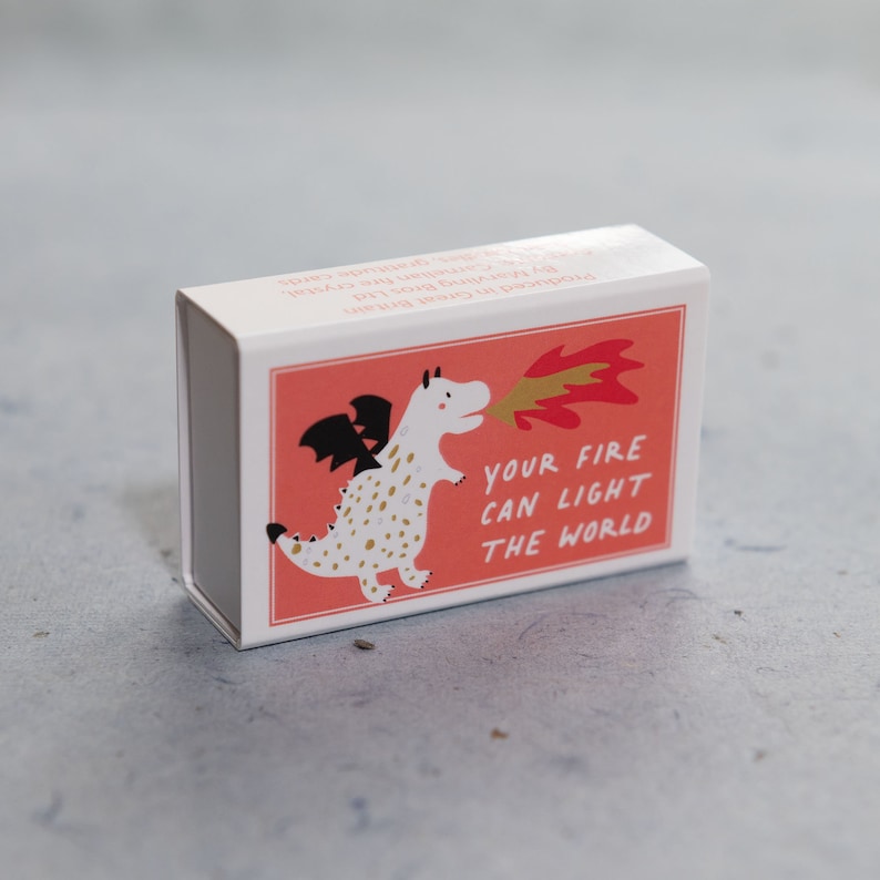 May include: A white matchbox with a red background and a cartoon white dragon with black wings and gold spots breathing fire. The text on the matchbox reads "Your fire can light the world."