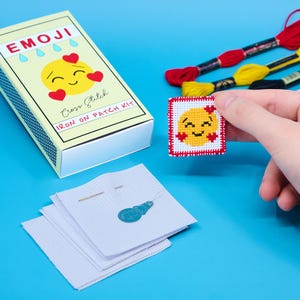 Op de afbeelding: Een gele emoji-kruissteekset met een lachend gezicht en hartjes. De set bevat een klein stukje Aida-doek, borduurgaren, een naald en een strijk-patch. De set is verpakt in een doos in de stijl van een luciferdoos met de woorden "Emoji Cross Stitch Iron On Patch Kit" op de voorkant.