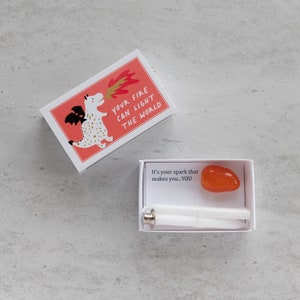 May include: A small white box with a red and white illustration of a dragon breathing fire. The text on the box reads "Your fire can light the world". The box is open and inside is a smaller white box with a small orange stone and two white sticks.