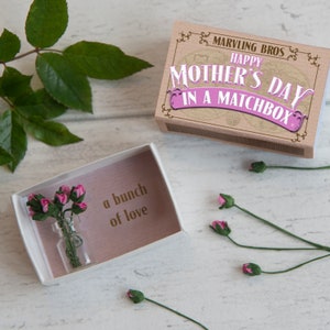 May include: A small white box with a pink interior containing a tiny glass bottle with a bouquet of pink flowers. The box has the text "a bunch of love" printed on the inside. The box is open and the flowers are visible. The box is sitting on a wooden surface. A larger box with the text "Happy Mother's Day in a Matchbox" is also visible in the image.