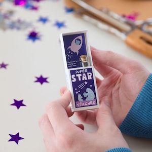 May include: A small white box with a purple and blue design. The box has a small glass vial inside and says "Super Star Teacher" on the front. The box is being held by a person's hands.