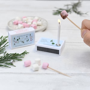 May include: A matchbox with a lit candle, toasting a marshmallow on a skewer. The matchbox has a blue and white design with the text "Happiness is roasting marshmallows together." Another matchbox with the text "Wishing you big Christmas adventures" is also visible.