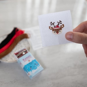 May include: A close-up of a completed cross-stitch project featuring a red-nosed reindeer wearing a Santa hat. The cross-stitch is on white Aida cloth and is framed by a white cardboard backing. The project is part of a craft kit that includes floss and instructions.
