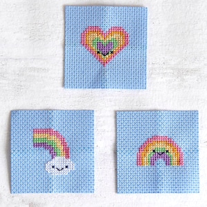 May include: Three small blue fabric squares with rainbow cross-stitch designs. The first square has a rainbow heart, the second has a rainbow with a white cloud, and the third has a rainbow.