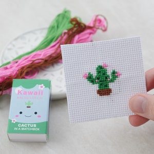 May include: A cross-stitch cactus design on white fabric, held in a hand, with green, pink, and brown embroidery floss in the background. A small box labeled "Kawaii Kross Stitch" with "Cactus in a Matchbox" is also visible.