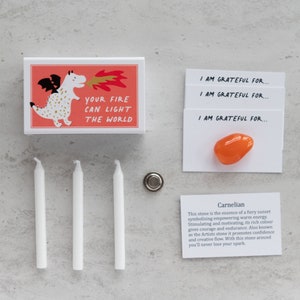 May include: A pink and white box with a cartoon dragon blowing fire. The box says "Your fire can light the world". Three white candles, a small metal disc, and a card with a carnelian stone and the text "I am grateful for..." are also shown. The card also includes a description of the carnelian stone.