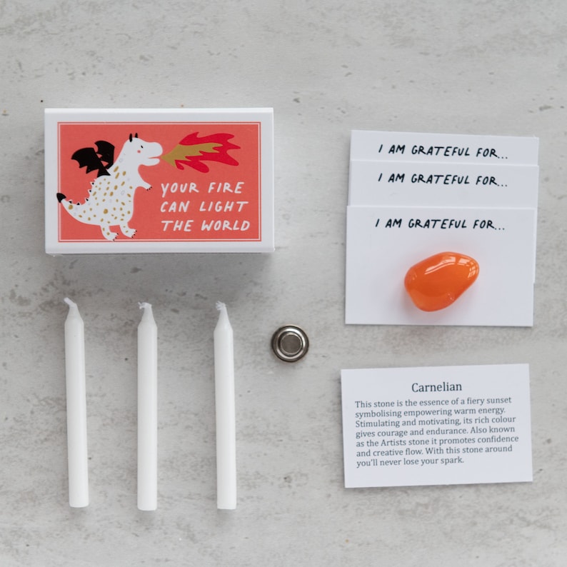 May include: A pink and white box with a cartoon dragon blowing fire. The box says "Your fire can light the world". Three white candles, a small metal disc, and a card with a carnelian stone and the text "I am grateful for..." are also shown. The card also includes a description of the carnelian stone.