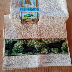 Könnte beinhalten: Zwei weiße Badetücher. Ein Handtuch hat einen Rand mit drei schwarzen Labrador-Retrievern in einem Feld mit weißen Blumen. Das andere Handtuch hat einen Rand mit Golfmotiven, darunter ein Teddybär-Golfer.