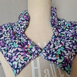 Könnte beinhalten: Ein Nackenwärmer aus Stoff mit einem floralen Muster in Violett-, Grün- und Weißtönen auf dunkelblauem Hintergrund. Der Wärmer ist mit Reis oder Leinsamen gefüllt und kann für therapeutische Zwecke erwärmt oder gekühlt werden.