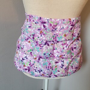 Könnte beinhalten: Ein rosa Stoffgürtel mit einem Blumendruck mit weißen, violetten und grünen Blättern. Der Gürtel ist gesteppt und fühlt sich weich und angenehm an.