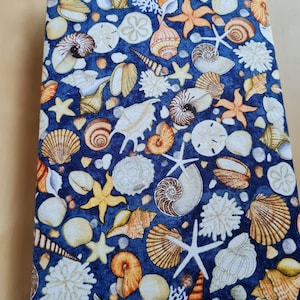 Könnte beinhalten: Ein blauer Stoffbuchumschlag mit einem Muschelmuster. Das Muster enthält verschiedene Muscheln, Seesterne und Korallen in Weiß-, Braun- und Orangetönen.