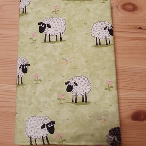 Könnte beinhalten: Ein rechteckiger, hellgrüner Stoffartikel mit einem sich wiederholenden Muster aus Cartoon-Schafen, rosa Blumen und Bienen. Die Schafe haben weiße Wolle, schwarze Gesichter und schwarze Beine. Der Hintergrund ist hellgrün mit einem dezenten, melierten Effekt.