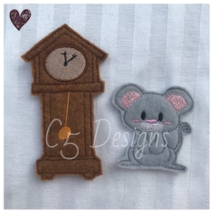Puede incluir: Un reloj de fieltro marrón con una esfera blanca y un péndulo negro. Un ratón de fieltro gris con orejas rosas y una nariz rosa se sienta junto al reloj.