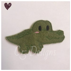 May include: A green felt alligator applique with black eyes and a pink mouth. The alligator is smiling and has a stitched outline.