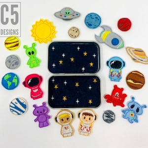 May include: A set of felt patches with a space theme. The patches are in various shapes and colors, including a sun, a spaceship, planets, and aliens. The patches are arranged on a white background with a blue and white tin box in the center.