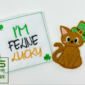 May include: A white felt square with green embroidery that reads "I'm Feline Lucky" and a green shamrock on each side. A brown felt cat wearing a green leprechaun hat with a gold buckle and a green shamrock is to the right of the square.