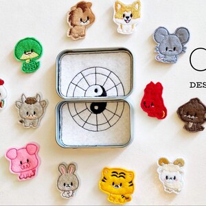 May include: A set of 12 embroidered iron-on patches featuring the Chinese zodiac animals. The patches are in a variety of colors and designs, and are arranged around a small tin box with a yin-yang symbol on the lid. The text "C5 DESIGNS" is visible in the upper right corner of the image.