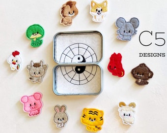 May include: A set of 12 embroidered iron-on patches featuring the Chinese zodiac animals. The patches are in a variety of colors and designs, and are arranged around a small tin box with a yin-yang symbol on the lid. The text "C5 DESIGNS" is visible in the upper right corner of the image.