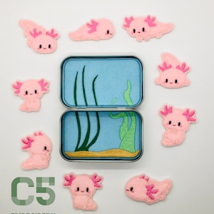 May include: Nine pink axolotl embroidery patches with black eyes and pink gills. The patches are arranged around a small blue and green tin with a sand bottom and green seaweed.