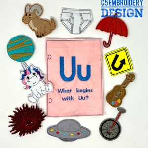 May include: A pink felt board with the letter "Uu" embroidered in blue. The board has the text "What begins with Uu?"  Surrounding the board are various felt patches, including a unicorn, a red umbrella, a yellow road sign, a grey spaceship, a red sea urchin, a brown ukulele, a grey unicycle, a white pair of underwear, a brown ram, and a blue and green planet.