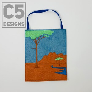 May include: A felt wall hanging with a blue sky, brown hills, and two green trees. The hanging has a blue loop at the top for hanging.