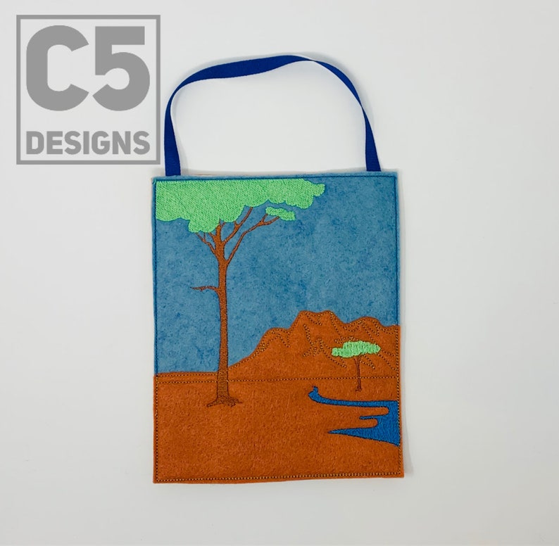 May include: A felt wall hanging with a blue sky, brown hills, and two green trees. The hanging has a blue loop at the top for hanging.