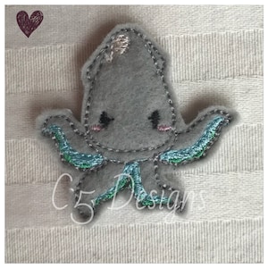 May include: A gray felt octopus with teal and silver embroidered details. The octopus has a cute, cartoon-like face with black eyes and pink cheeks.
