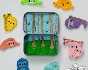 May include: A set of felt sloth finger puppets in various colors, including green, yellow, purple, pink, and blue. The puppets are arranged around a small metal tin with a felt scene of a jungle with trees and vines. The tin is open and the scene is visible. The text "C5 EMBROIDERY DESIGN" is visible in the bottom right corner of the image.