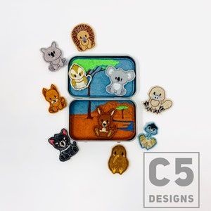 May include: A set of felt animal finger puppets in a tin box. The puppets include a koala, kangaroo, echidna, platypus, kookaburra, Tasmanian devil, and lion. The tin box has a scene of a kangaroo in a desert landscape.