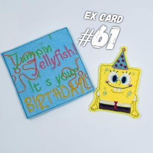 Puede incluir: Una tarjeta de fieltro azul con el texto "Jumpin Jellyfish! It's your Birthday!" y un personaje de dibujos animados bordado en amarillo que lleva un gorro de fiesta azul y blanco.