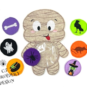 Puede incluir: Una momia de fieltro con un bolsillo transparente, rodeada de círculos de fieltro con diseños de Halloween. Los círculos presentan un hueso, un fantasma, una araña, una bruja, un cuervo y una rata. El texto "CS EMBROIDERY DESIGN" está en la parte inferior.