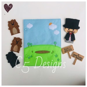 May include: Felt finger puppets of a groundhog and a man in a top hat. The puppets are on a felt background with a green field, a blue sky, and clouds. There are two signs that say "Early Spring" and "6 More Weeks of Winter".