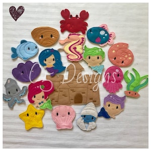 May include: A set of felt ocean-themed finger puppets, including a mermaid, a crab, a seahorse, a starfish, a seashell, a fish, an octopus, a jellyfish, and seaweed. The puppets are arranged around a felt sandcastle.