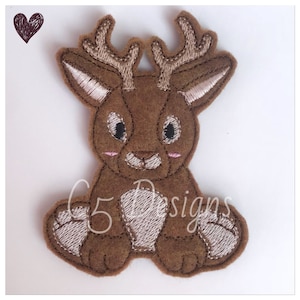 May include: A brown felt reindeer applique with a white face and large eyes. The reindeer is sitting with its legs crossed and has antlers.