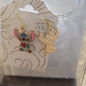 Puede incluir: Un par de pendientes colgantes dorados con el personaje Stitch de la pel&iacute;cula de animaci&oacute;n Lilo & Stitch. Un pendiente muestra a Stitch con cuerpo azul, orejas rosas y un coraz&oacute;n rojo. El otro pendiente es una silueta dorada.