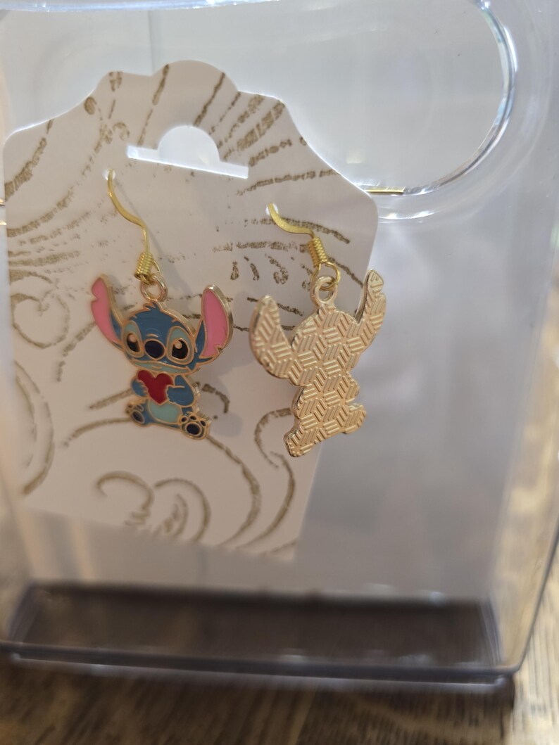Puede incluir: Un par de pendientes colgantes dorados con el personaje Stitch de la pel&iacute;cula de animaci&oacute;n Lilo & Stitch. Un pendiente muestra a Stitch con cuerpo azul, orejas rosas y un coraz&oacute;n rojo. El otro pendiente es una silueta dorada.