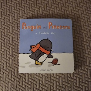 May include: A children's book cover with a blue background and the title "Penguin and Pinecone, a friendship story". The cover features a cartoon penguin wearing a red and orange scarf, standing on snow with a pinecone in front of it.