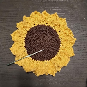 Può includere: Un sottobicchiere a forma di girasole fatto a mano all'uncinetto, con un centro marrone e petali gialli. Un uncinetto argentato è appoggiato sul sottobicchiere. Il sottobicchiere è progettato per assomigliare a un girasole.