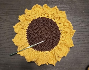 May include: A handmade crochet sunflower coaster with a brown center and yellow petals. A silver crochet hook rests on the coaster. The coaster is designed to resemble a sunflower.