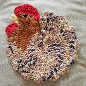 May include: A handmade crochet trivet in the shape of a rooster. The trivet features a red comb and wattle, brown body, and a multi-colored tail with shades of brown, pink, and yellow. The trivet is designed to protect surfaces from hot items.