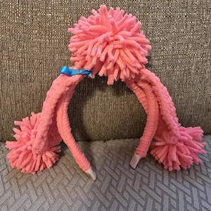 May include: A pink headband featuring a large, textured pom-pom at the top and two smaller pom-poms at the ends of the band. The headband is covered in soft, fuzzy yarn and includes a small, bright blue ribbon bow attached near the top pom-pom.