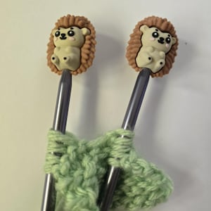 May include: Two silver-colored knitting needles featuring decorative toppers shaped like small, light brown hedgehogs with black eyes and pink cheeks. The needles are holding a piece of light green knitted yarn fabric in a simple stitch pattern.